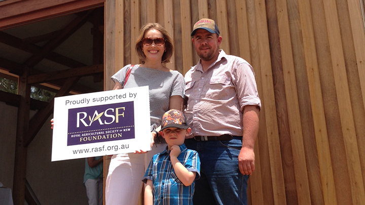 Family of three standing outside a timber building, holding a sign supporting the RAS Foundation.