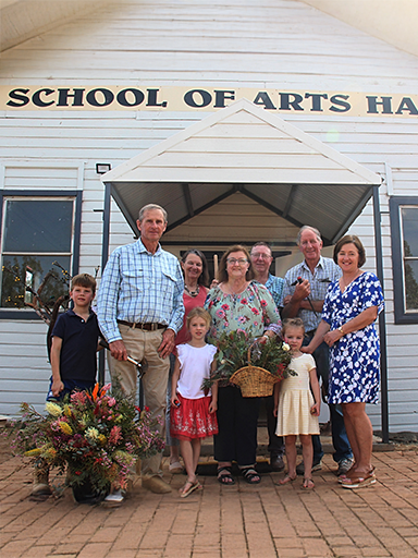 A group of adults and children standing in front of a white building, smiling and holding flowers and baskets.