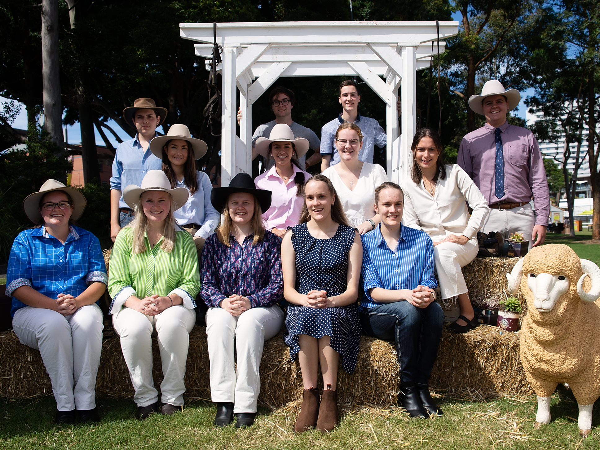 The RAS Rural Scholaship recipients, smiling and wearing rural attire and hats, seated on hay bales outdoors with a ram statue.