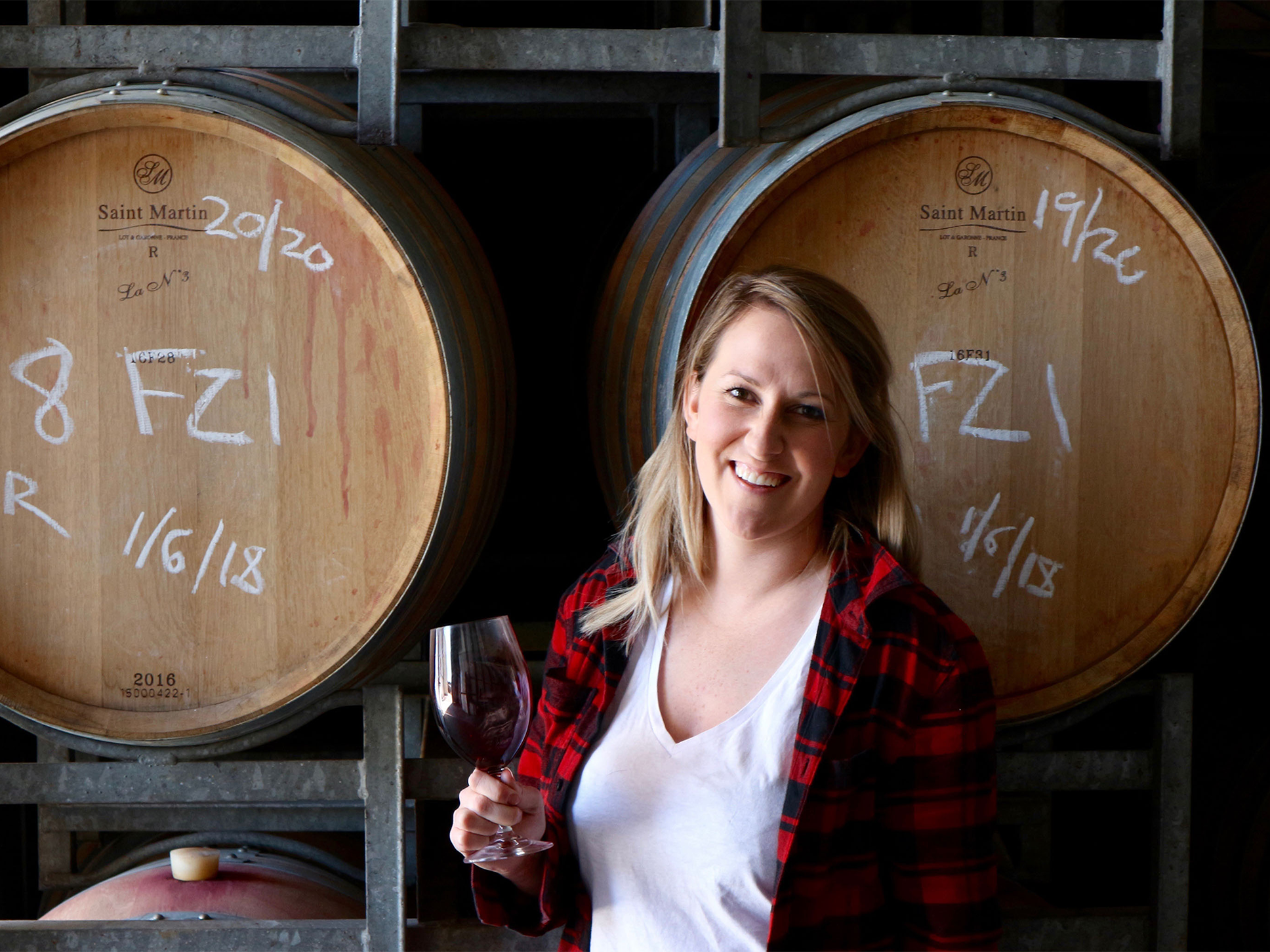 A Sydney Royal Wine Scholarship recipient holding a glass of red wine, smiling and standing in front of wine barrels in a cellar.
