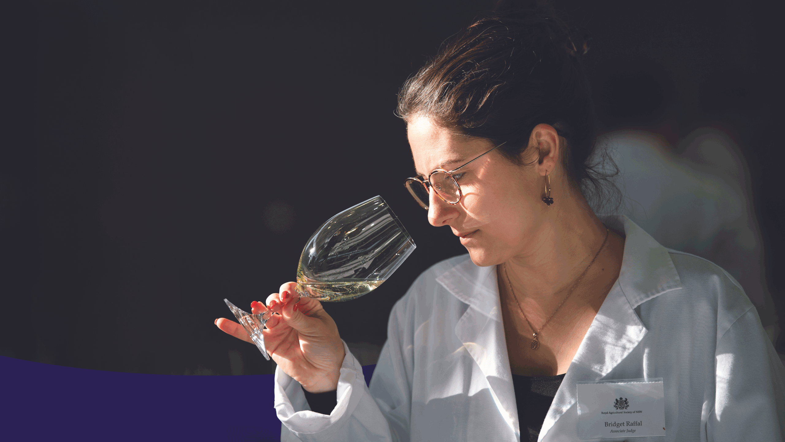 A judge in a white coat and glasses  carefully inspecting a glass of white wine at the Sydney Royal Wine Competition.