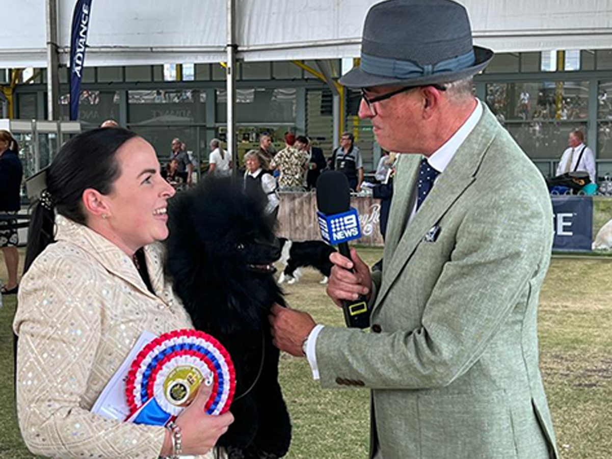 A competition prize winner with a black dog is interviewed by a reporter holding a microphone.