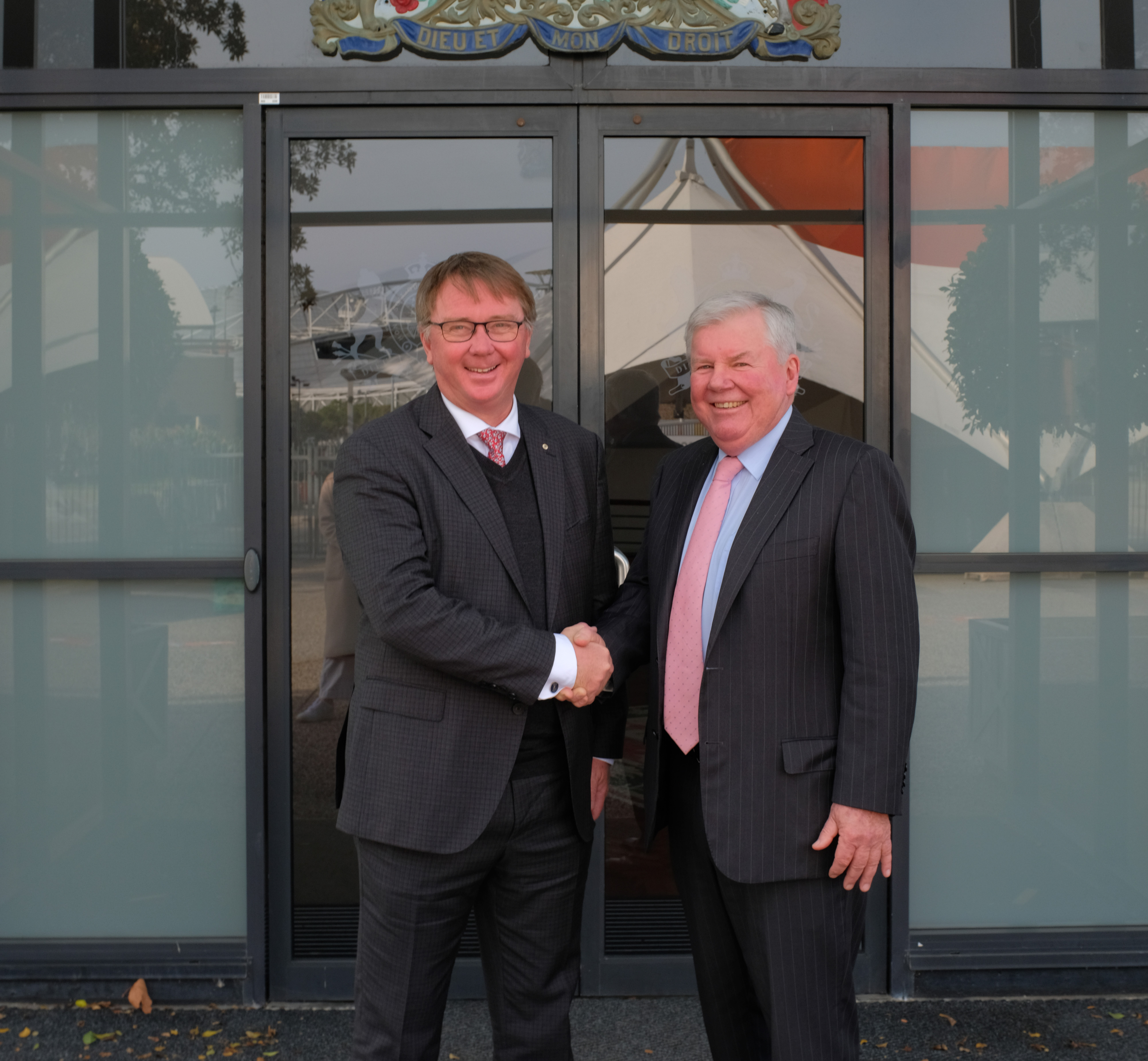 Two men in suits shake hands outside a building, smiling at the camera.