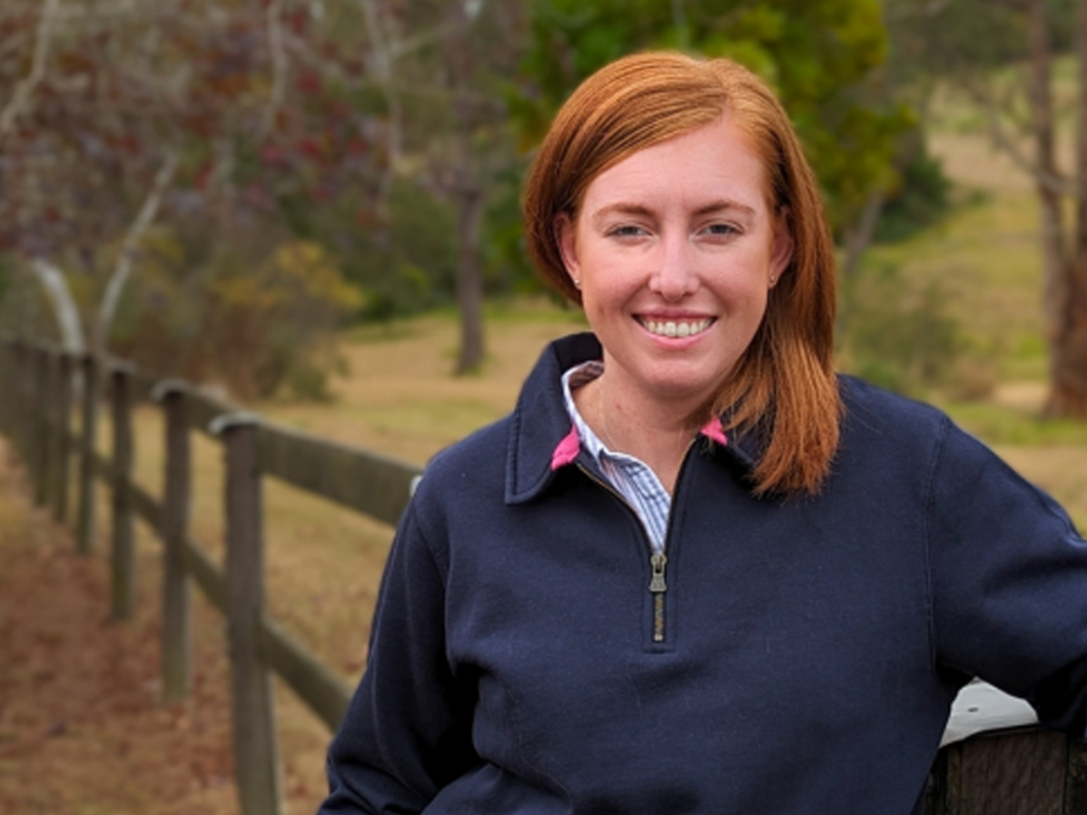RAS Youth Group Member Courtney Nelson with red hair, wearing a navy jumper and smiling  while leaning on a wooden fence.