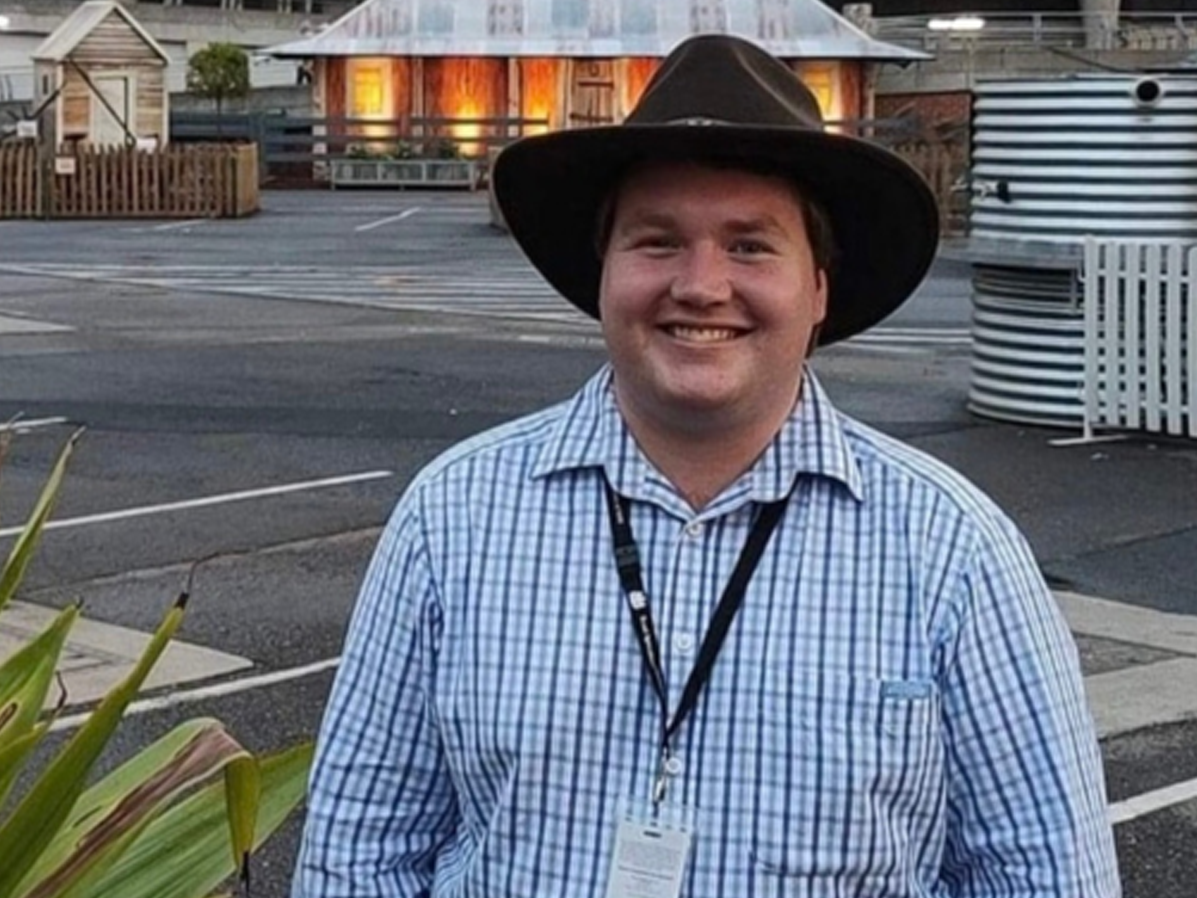 RAS Youth Group member Michael Roffe in a checked shirt and wide-brimmed hat stands outdoors near rural-style buildings.