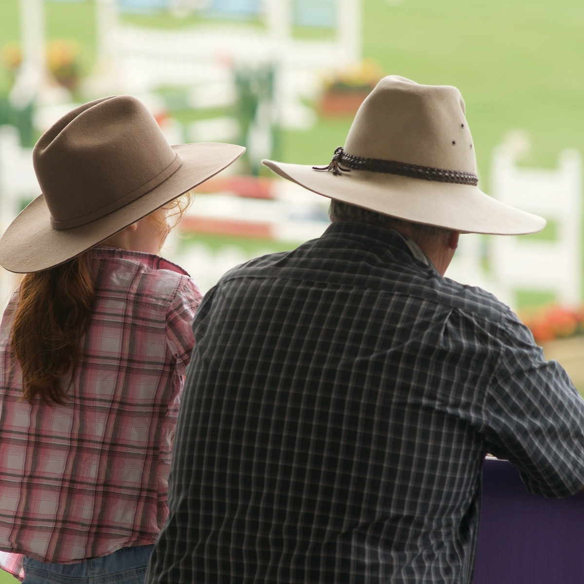 A child and adult wearing wide-brimmed hats, watching an event at an outdoor arena.